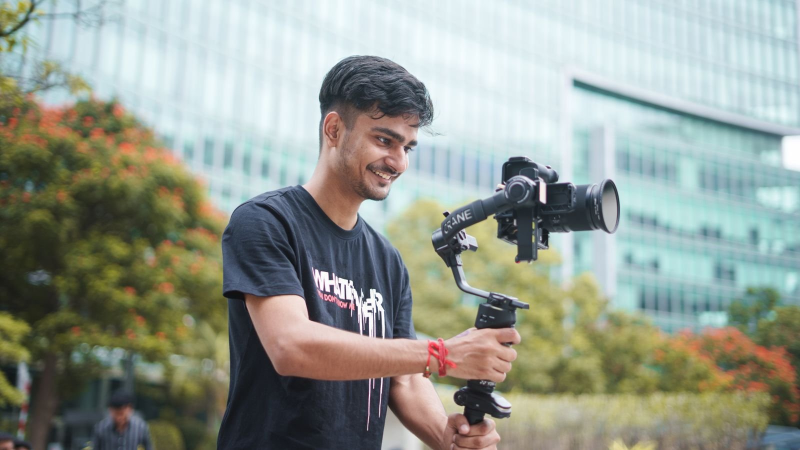 Smiling young videographer using a gimbal outdoors. Urban setting and greenery in the background.