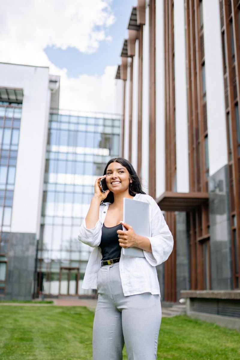 Confident woman holding laptop, talking on phone outside an office building.